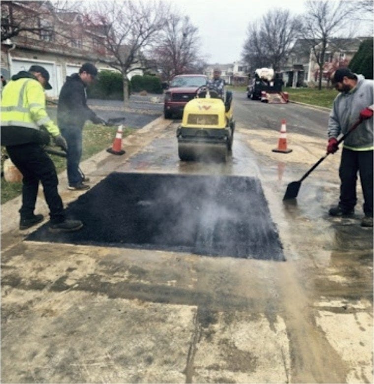 Three of the crew working a hot patch, steam still coming off it, roller waiting behind.