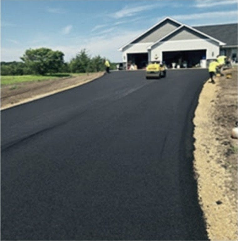 A finished black drive running out from a house between mown grass and a fresh sand shoulder.