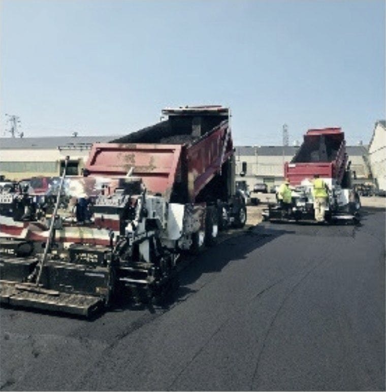 Paver and trucks working a commercial lot, crew alongside.