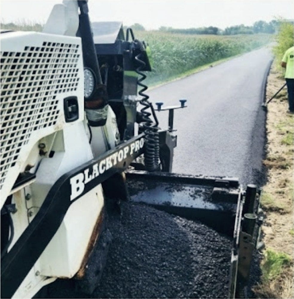 The paver laying a fresh mat beside a standing crop, the Blacktop Pro&rsquo;s name on the machine.