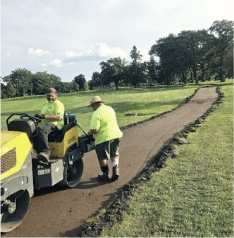 A roller and two of the crew working a new path across mown turf, mature trees behind.
